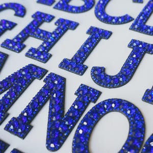 May include: Close-up of several blue rhinestone-covered letters, including H, I, J, N, and O, against a white background. The letters are dark blue with a black outline, and the rhinestones sparkle. The letters are likely for crafts or personalisation.
