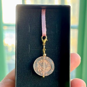 以下が含まれることがあります： 金色の留め金とピンクのリボンから吊り下げられた、花柄のデザインが施された丸い薄ピンク色のチャーム。 チャームは黒い箱に入っており、ギフトやジュエリーを連想させます。 背景はぼやけています。