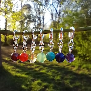 Op de afbeelding: Zeven zilveren bedels met elk een kleine ronde edelsteen. De edelstenen zijn rood, oranje, geel, groen, blauw, indigo en violet en vertegenwoordigen de zeven chakra's.