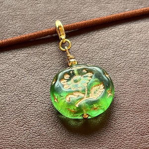 Könnte beinhalten: Runder, grüner Glasanhänger mit goldenem Blumendesign und goldenen Akzenten. Der Anhänger hängt an einem goldenen Verschluss und einer braunen Kordel. Der Hintergrund ist eine strukturierte, braune Oberfläche.