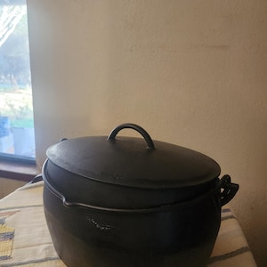 May include: A black cast iron Dutch oven with a lid and handle. The pot has a rounded shape and two side handles. The lid has a handle on top. The pot is sitting on a patterned tablecloth.