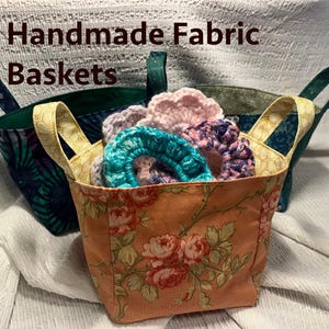 May include: Three fabric baskets with floral patterns. The baskets are filled with crocheted items in shades of pink, blue, and purple. The text "Handmade Fabric Baskets" is displayed above the baskets.