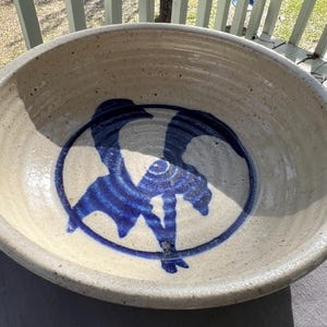 May include: A large, round stoneware bowl with a speckled, off-white exterior. The interior features a blue abstract design within a blue circle. The bowl has a wide rim and a deep interior, suitable for serving or display.