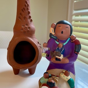 May include: Two ceramic figurines. One is a tall, brown, chimney-shaped piece with decorative cutouts. The other is a colorful figure of a person in a purple robe, surrounded by several small figures. The figures appear to be a family.