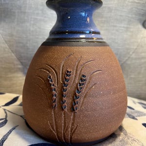 May include: A handcrafted ceramic vase with a brown body and a blue neck. The vase features an embossed wheat stalk design. The vase is a decorative item for the home.