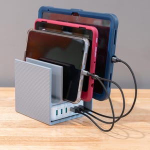 May include: A light gray multi-device charging station with a ribbed exterior texture. The stand holds three tablets in protective cases, colored blue, pink, and clear, each connected to a white multi-port USB charger base with black cables.