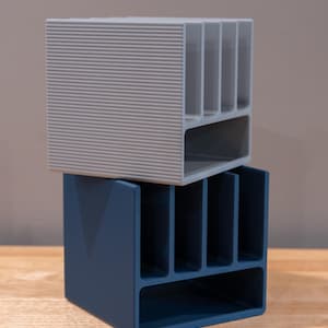 May include: A light gray desk organizer with a ribbed exterior texture stacked on top of a solid dark blue desk organizer. Both organizers feature four vertical slots and one horizontal rectangular compartment for storage and organization.