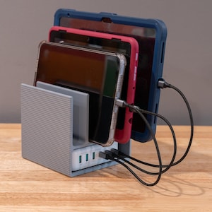 May include: A gray plastic charging station with a ribbed exterior design, featuring three vertical slots holding a tablet in a clear case, a tablet in a pink case, and a tablet in a blue case, all connected to a white multi-port USB charger.
