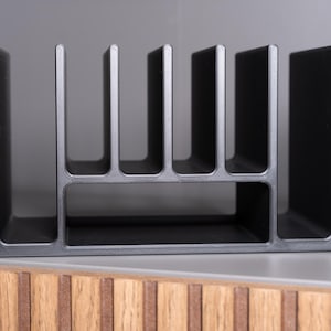 May include: A matte black plastic organizer featuring a series of vertical dividers creating multiple slots for storage. The structure has a rectangular base with a lower horizontal compartment and six upright fins extending upwards from the frame.