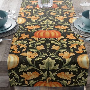 May include: A decorative table runner featuring a repeating pattern of orange pumpkins and autumn leaves on a black background. The runner is set on a wooden table, with place settings and a vase of pink flowers visible.