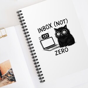Puede incluir: Cuaderno blanco con espiral con el texto "INBOX (NOT) ZERO" y una ilustración de un gato negro con gafas. El cuaderno también presenta un gráfico de computadora portátil con una notificación por correo electrónico. También se ve un clip de papel dorado y gafas de sol.