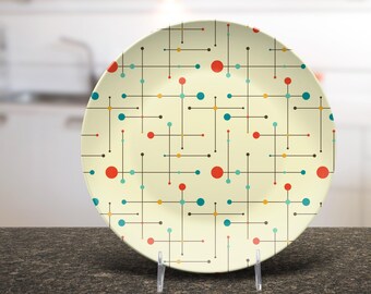 May include: A round decorative plate with a cream background. It has a geometric pattern of intersecting black lines, red, teal, yellow, and black dots. The plate is displayed on a clear stand.