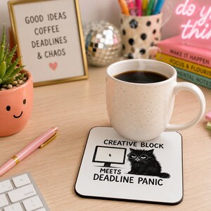 Pode incluir: Uma caneca branca salpicada cheia de café está em cima de um porta-copos quadrado com um gráfico de gato preto e o texto "CREATIVE BLOCK MEETS DEADLINE PANIC". Uma placa emoldurada diz "GOOD IDEAS COFFEE DEADLINES & CHAOS".