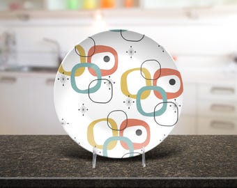May include: A white ceramic plate with a retro design. The plate features a pattern of overlapping squares and circles in teal, orange, and yellow, with black accents and starburst details. The plate is displayed on a clear stand.