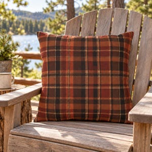 May include: A square throw pillow featuring a plaid pattern in shades of burnt orange, deep brown, and black. The fabric has a woven texture with intersecting horizontal and vertical lines forming a classic grid design across the entire surface.