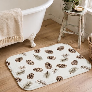 Op de afbeelding: Een rechthoekige badmat met een witte achtergrond voorzien van een herhalend patroon van bruine dennenappels en groene dennennaalden. De mat heeft afgeronde hoeken en een zachte textuur, geplaatst op een lichte houten vloer in een badkamer.