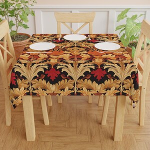 May include: A rectangular tablecloth featuring a repeating pattern of gold, red, and orange stylized leaves on a black backdrop. The tablecloth is laid on a wooden table with four chairs, and three white plates are visible.
