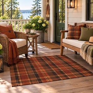 May include: A rectangular area rug featuring a classic plaid pattern in shades of burnt orange, dark brown, and black. The rug is positioned on a wooden porch floor, surrounded by wicker and wooden furniture, potted plants, and a lit lantern.