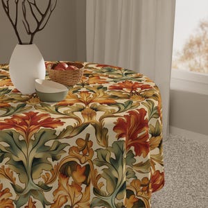Puede incluir: Un mantel redondo con fondo crema y un patrón repetido de hojas estilizadas en tonos naranja, verde y dorado. Un jarrón blanco con ramas, una pequeña cesta de manzanas y un cuenco verde y blanco están sobre la mesa.