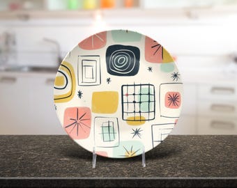 May include: A decorative plate with a mid-century modern design. The plate has a cream background with colourful geometric shapes in pink, yellow, blue, and black. The plate is displayed on a clear stand.