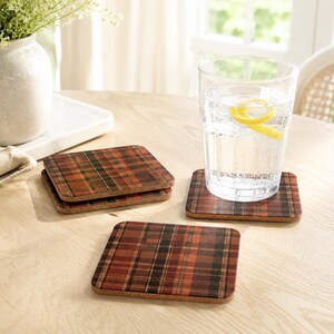 May include: A set of three square coasters featuring a dark red, brown, and black plaid pattern. The coasters have a cork base and a smooth top surface. One coaster supports a clear glass filled with water and a yellow lemon peel twist.