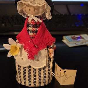 May include: A handmade fabric doll dressed in traditional attire. The doll wears a black hat, red shawl, and a striped apron. It holds a daffodil and has a tag that says "Sea".
