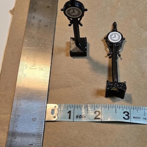 May include: Two miniature black street clocks with white clock faces, standing on a brown surface. A metal ruler and a measuring tape are also present. The measuring tape shows approximately 3 inches.