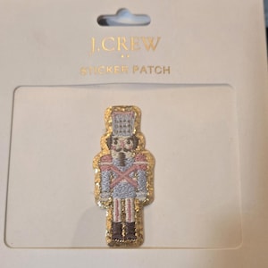 May include: A J.Crew sticker patch featuring a gold-trimmed nutcracker soldier in a light blue uniform with pink and gold accents. The patch is on a white background with the J.Crew logo.