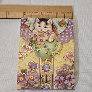 May include: A decorative greeting card featuring a kitten in a teacup surrounded by purple flowers and butterflies. The card has a gold and purple floral design. The ruler shows a measurement of approximately 10 cm.
