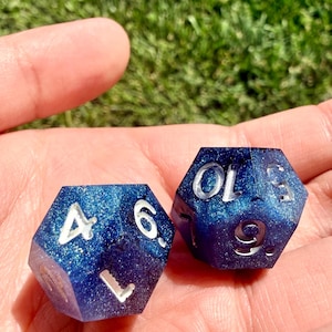 May include: Two multi-sided polyhedral dice with a deep blue and purple glitter finish. The dice feature engraved numbers filled with silver paint, including the visible digits 4, 6, 9, and 10. The dice have a smooth, polished surface texture.