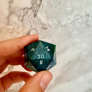 May include: A handmade, twenty-sided die with a teal and dark blue marbled effect. The numbers are white and clearly visible. This polyhedral die is perfect for tabletop role-playing games.