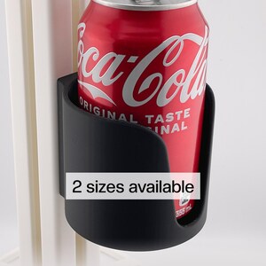 Könnte beinhalten: Ein schwarzer Getränkehalter mit einer roten Coca-Cola-Dose. Die Dose hat weißen Text und fasst 355 ml. Der Text "2 Größen verfügbar" ist auf dem Getränkehalter zu sehen.