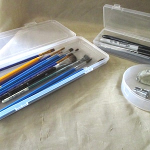 May include: Three clear plastic containers with lids. The largest container holds a variety of paintbrushes and pencils. The medium-sized container holds a black marker and a silver Sharpie. The smallest container holds a pencil sharpener and a gray eraser.