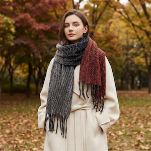May include: A long, rectangular scarf featuring a color-blocked design with a dark gray, textured herringbone pattern on one side and a solid, speckled rust-red section on the other, finished with long, dark fringe tassels at both ends.