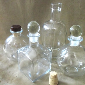 May include: A collection of clear glass bottles and jars in various shapes and sizes. Some bottles have glass stoppers, while others have cork stoppers. The bottles are arranged on a neutral-toned surface, showcasing their transparency and design.
