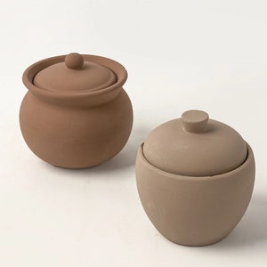 May include: Two small, lidded terracotta pots. One pot is a rounded shape with a darker brown color, and the other is a more oval shape with a lighter tan color. Both pots have a small knob on top of the lid.