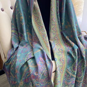 May include: A teal shawl with a paisley pattern and a colorful border. The shawl is draped over a chair and has a fringe along the bottom edge. The design includes shades of teal, purple, yellow, and pink.