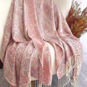 Peut inclure: Une couverture rose à motifs cachemire avec des franges crème est drapée sur une chaise blanche. La couverture présente un motif détaillé dans les tons de rose, pêche et crème. Un élément de décoration intérieure.