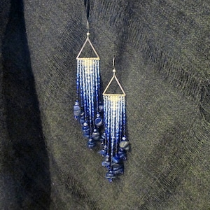 May include: A pair of handmade earrings with a geometric design. The earrings feature a silver triangle top with beaded fringe in shades of blue and white. Dark blue stone accents are at the bottom of the fringe.
