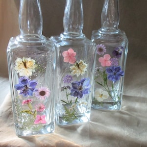 May include: Three clear glass bottles with a square shape and a textured surface. Each bottle contains pressed flowers in shades of pink, white, blue, and green.