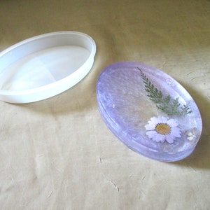 Puede incluir: Una bandeja ovalada de resina de color lila claro con un diseño de flores y helechos prensados. La bandeja se muestra con su molde de silicona transparente.