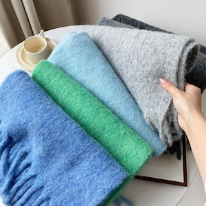 May include: A stack of folded scarves in various colors including bright blue, vibrant green, light blue, light gray, and dark charcoal gray. Each scarf features a soft, fuzzy, brushed texture with visible fibers and fringe details along the edges.