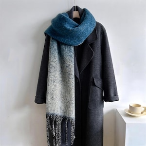 May include: A dark charcoal gray coat with a long, thick scarf featuring a gradient pattern that transitions from a deep blue at the top to a light gray at the bottom, finished with long, twisted fringe tassels at the hem.
