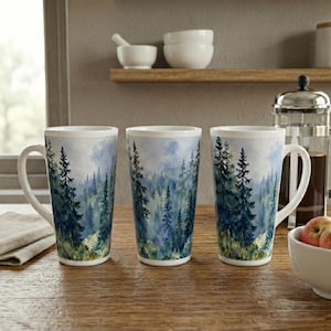May include: Three tall ceramic mugs featuring a watercolor-style forest landscape with dark green pine trees against a soft blue and white sky. Each mug has a white interior and a sturdy handle, arranged on a wooden surface.