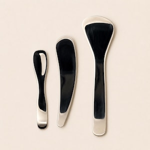 May include: A set of three kitchen utensils with polished silver-toned metal edges and smooth, dark black centers. The collection includes a small narrow spoon, a medium-sized curved spreader, and a larger, wider spatula with a flared head.