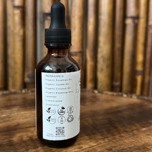 May include: A brown glass bottle with a black dropper, filled with oil. The label lists ingredients like Organic Rosehips Oil, Lavender, and Chamomile. The bottle has several icons and the text "Expires Sept 2027 discover more."