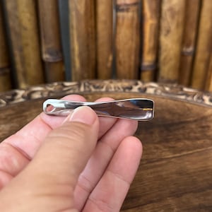 Könnte beinhalten: Ein polierter silberfarbener Anhänger mit geschwungenem, länglichem Design. Der Anhänger hat an einem Ende ein kleines Loch und wird in der Hand einer Person gehalten. Der Hintergrund zeigt Bambusstängel und eine Holzoberfläche.