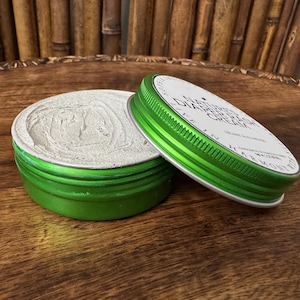 Könnte beinhalten: Geöffnete grüne Dose mit Windelausschlagcreme, der Deckel leicht geöffnet. Der Deckel hat weißen Text auf weißem Hintergrund. Die Creme in der Dose ist hellgrau. Die Dose steht auf einer Holzoberfläche.