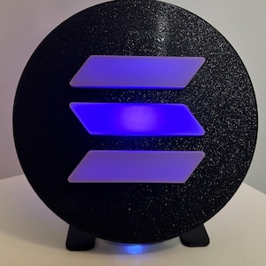 May include: A black circular light with a white and blue three-dimensional logo in the center. The light is on a black stand.