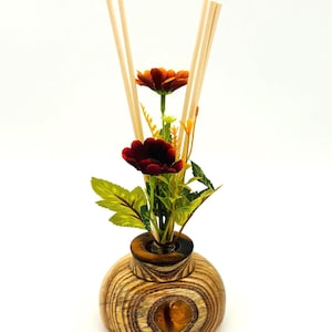 May include: A decorative reed diffuser with a wooden base and a glass neck. The diffuser holds artificial flowers and reeds. The flowers are red and orange, and the reeds are light brown. The wooden base has a natural wood grain pattern.